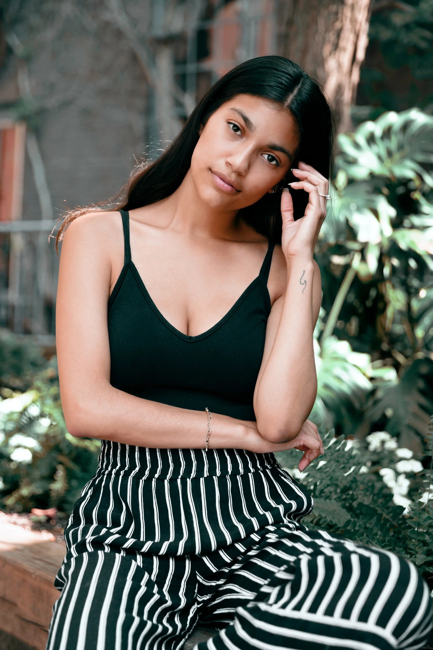 Woman in a black top and striped pants sitting outdoors with greenery in the background